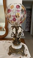 Full view of lamp showing the glass globe with pink, amber, and gold circular accents, gold-tone cherubs on white marble base, and crystal hanging prisms.