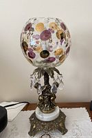 Full view of the lamp showing the glass globe with colored circular accents, gold cherub figures, crystal prisms hanging below the globe, white marble mid-base, and ornate metal bottom base.