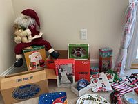 Large stuffed wired Santa Claus sitting with various boxes and tins, including a Santa cookie jar box in front.