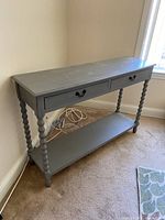 Gray console table against a wall showing top surface, two drawers with decorative black handles, spiral legs, and bottom shelf. Minor wear visible.
