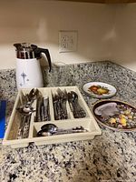 Photo showing vintage Corning Ware percolator with blue starburst pattern, stainless steel silverware in compartmentalized tray, small clear glass pitcher, and two decorative fruit-themed plates on granite countertop.