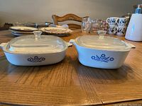 View of two Corning Ware ceramic casserole dishes with covers from a slightly diagonal angle on wooden table.