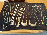 Photo showing a collection of 14 mostly vintage costume jewelry necklaces laid on a black cloth, including faux pearl strands, beaded necklaces, and a statement bib necklace along with a pair of clip-on earrings.