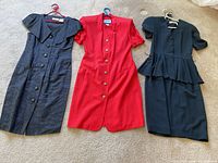 Full view of three dresses on hangers laid out on floor: black dress with large collar and buttons, red dress with gold buttons, black dress with peplum waist and buttons, all in good condition.