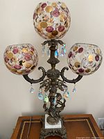 Full view of the lamp showing three glass globes with gold and purple circular accents, metal base, and white marble-like middle base with cherub figure.