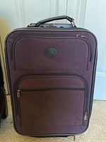 Front view of the medium purple luggage piece with fabric front, outer zippered pockets, and logo patch.