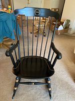 Front view of Nichols & Stone black wooden rocking chair with gold trim and vertical spindle backrest, showing commemorative plaque at center of back rail.