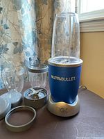 Nutribullet Magic Bullet blender with blue base, blending cup on top, blade assembly, extra cups, lid, and ring displayed on dark surface near window with floral curtain.
