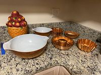 Two photos showing a set of kitchen collectibles: two Pyrex mixing bowls, a ceramic apple basket cookie jar, and five copper-tone decorative baking molds on a marble counter.