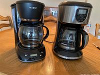 Two drip coffee makers side by side on a wooden table, showing front view with glass carafes, control panels, and brand names visible.