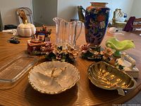 Photo of assorted ceramic and glassware items on table including Fenton custard dish, lusterware vase, clear glass pitcher, music box with mice, Capodimonte candle holders, and other serving dishes