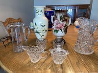 Front view of the lot showing two large ceramic vases with floral paintings, three clear cut glass pitchers, sugar bowl, and creamer on a wooden table.