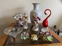 Wide shot of Victorian style porcelain figures, decorative vase, glass swan sculpture, glass pedestal candy dish, and floral porcelain candy dishes arranged on a glass table.