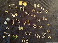 Photo showing the full assortment of 30 pairs of vintage costume earrings, including studs, wired styles, and different shapes and colors, along with the sweater clip and pendant on black fabric.