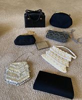 Overview of seven vintage purses and a metallic wallet arranged on carpet: includes black textured handbag, black fabric clutch, black crocheted purse with chain, silver beaded wallet, silver gray beaded purse, white sequined purse, and flat black clutch.