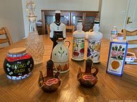 Photo showing entire collection of decorative bottles and decanters on a wooden table with chairs in background. Items include crystal decanter, ceramic bottles with fruit and floral designs, novelty 'Casino Cash' jar, and oil and vinegar ceramic cruets.