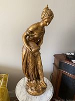 Full view of tall gold painted woman statue holding an urn, standing on round base.