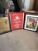 Three framed prints arranged on floor, held by person. Left print is vintage newspaper page, center is red Keep Calm and Carry On poster, right is British war savings propaganda poster.