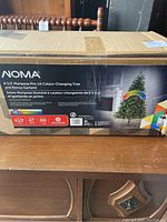 Box packaging displaying the NOMA brand 6.5 foot Mariposa pre-lit colour-changing tree and bonus garland information, with a photo of the tree on the box.