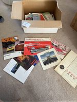Photo of assorted car brochures inside and outside a cardboard box showing multiple brochures with vintage car images including a 1958 De Soto, 1980 Pontiac, VW Golf Cabriolet and others.