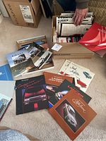 Multiple vintage car brochures from different decades spread on carpet and in boxes, featuring various manufacturers and models.
