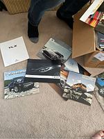 Multiple car brochures laid out on carpet floor showing Toyota Tacoma, Subaru Impreza WRX STI, Acura TLX with a visible arm and legs above them.