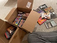 Photo showing multiple CDs in jewel cases and cassette tapes in cardboard box and scattered on carpet, featuring various music artists.