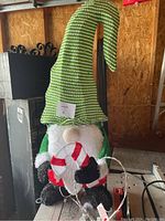 Front angle of the Christmas gnome showing its green and white striped tall hat, white beard, black gloves and boots, and holding a candy cane.