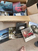 Box filled with GM car brochures with some brochures laid out on the floor: Cadillac CTS, Chevrolet H2, Pontiac Solstice, Saab 93, and Hummer H2 visible.