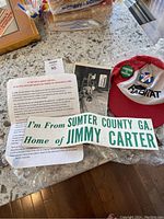 Image of red and white Habitat ball cap with three Jimmy Carter pins attached to front panel and a Jimmy Carter related bumper sticker and informational insert on the table.