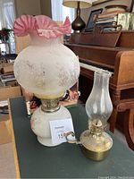 Photo showing white ceramic oil lamp with etched floral glass globe and pink ruffled top alongside smaller brass oil lamp with glass chimney and handle.