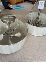 Two pendant light fixtures placed on carpet floor. Each has a round white drum shade about 18 inches diameter, metal frames with multiple arms holding light sockets visible inside the shades. One fixture is hanging from chain and wired for ceiling mounting.