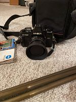 Photo showing Minolta X-700 camera with lens and attached lens hood sitting in front of black carrying case, with part of a tripod visible.