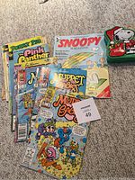 Photo showing a group of eight assorted vintage comics, including Muppet Babies, Snoopy Magazine, Porky Pig, and The Pink Panther, along with a small collectible Snoopy tin box.