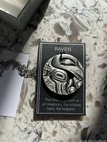 Front view of a round pewter pendant with a raven design placed on a card with descriptive text.