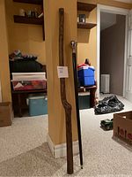 Two walking sticks leaning against a wall: a natural carved wood stick and a black wooden stick with metal top handle.