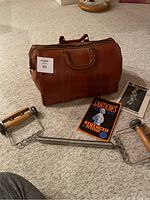 Photo of vintage brown leather duffel bag with two workout books, a hand grip strengthener, and a metal bar grip exercise tool on carpeted floor.