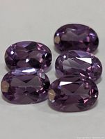 Close-up of 5 oval cut amethyst gemstones placed on a flat white surface showing the color and facets.