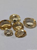Group of six faceted oval genuine citrine gemstones arranged on a white background, showing their size and color variations.