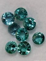 Close up of seven round cut green stones showing natural inclusions consistent with emeralds.