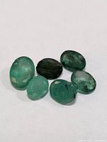 Photo showing six loose genuine emerald gemstones of varying green shades and oval shapes with natural inclusions.