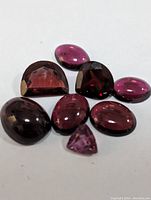 Seven garnet gemstones on a white background, showcasing a variety of cuts including cabochon and faceted with colors from dark red to reddish pink.