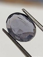 Close-up image of oval faceted iolite gemstone showing internal inclusions, translucent bluish-gray color, held by tweezers.