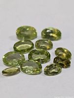 Collection of 11 faceted peridot gemstones arranged on a white surface showing different cuts and green-yellow shades.