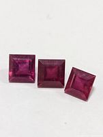 Photo of three square cut ruby gemstones placed side by side on white background, showing color and cut.