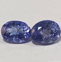 Close-up photo of two oval cut tanzanite gemstones showing blue-violet color and surface inclusions and damage.