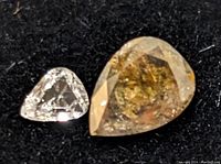 Close-up image showing two loose diamonds, the larger pear shaped salt and pepper diamond with visible inclusions and the smaller clear triangular diamond, both on a black surface.