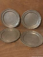 All four pewter plates arranged on wooden surface, showing overall size and shape with decorative scalloped edges