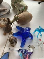 Overview of various small animal figurines made from glass, ceramic, and metal including blue starfish, glass fish, ceramic dog, and cracked ceramic egg with green animal head