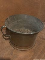 Heavy antique copper vessel with a single handle and a perforated bottom, shown from an angle to display the handle and shape.